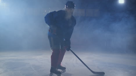 Man-hockey-player-with-a-puck-on-the-ice-in-hockey-form-leaves-with-a-stick-in-his-hands-out-of-the-darkness-and-looks-straight-into-the-camera